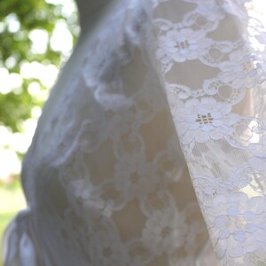 May include: A white lace garment featuring long sleeves with a detailed floral pattern. The fabric is semi-sheer, showcasing an intricate openwork design throughout the material. The garment is displayed on a form, highlighting the texture.