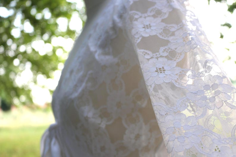 May include: A white lace garment featuring long sleeves with a detailed floral pattern. The fabric is semi-sheer, showcasing an intricate openwork design throughout the material. The garment is displayed on a form, highlighting the texture.