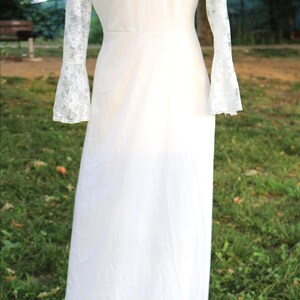 May include: A floor-length white dress featuring long sleeves with lace detailing at the cuffs. The garment has a fitted bodice and a flowing skirt, presented on a mannequin. The design includes a high neckline and a solid white fabric body.