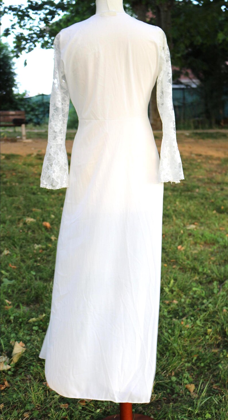 May include: A floor-length white dress featuring long sleeves with lace detailing at the cuffs. The garment has a fitted bodice and a flowing skirt, presented on a mannequin. The design includes a high neckline and a solid white fabric body.