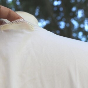 May include: A white garment featuring a delicate lace trim along the neckline and a small, rectangular fabric tag attached near the collar. The fabric appears smooth and lightweight, with a soft texture visible across the surface of the material.