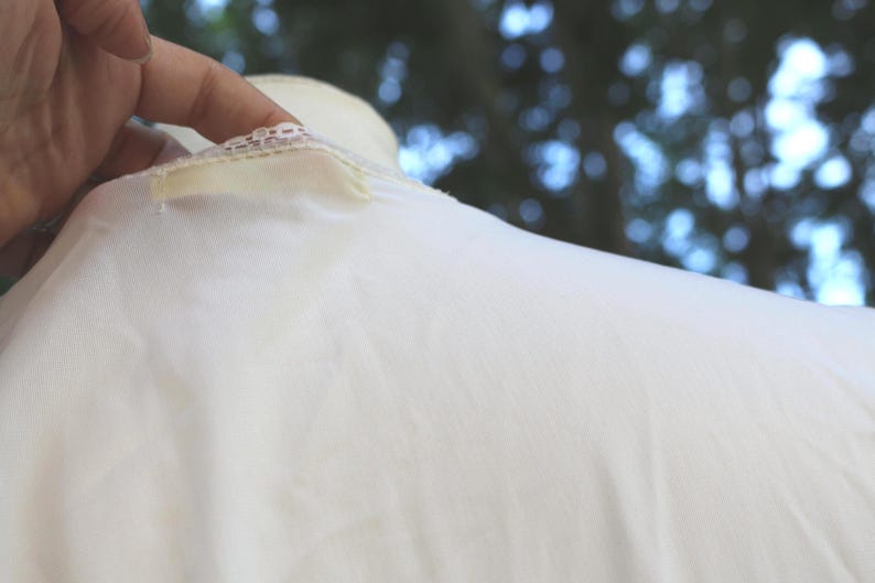 May include: A white garment featuring a delicate lace trim along the neckline and a small, rectangular fabric tag attached near the collar. The fabric appears smooth and lightweight, with a soft texture visible across the surface of the material.