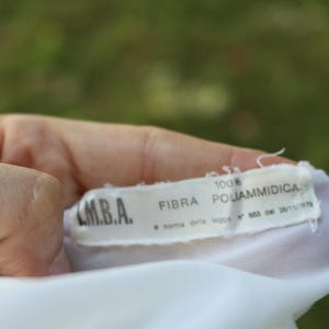 May include: A white fabric garment tag with black text reading M.B.A., 100% FIBRA POLIAMMIDICA, and a norma della legge N 883 del 26/11/1973. The rectangular label is stitched onto a white textile material with frayed edges at the top and bottom.