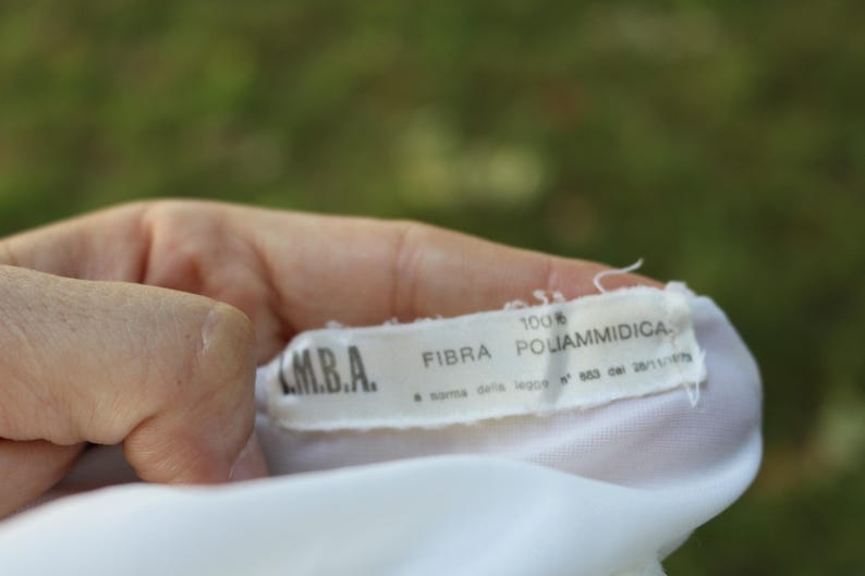 May include: A white fabric garment tag with black text reading M.B.A., 100% FIBRA POLIAMMIDICA, and a norma della legge N 883 del 26/11/1973. The rectangular label is stitched onto a white textile material with frayed edges at the top and bottom.