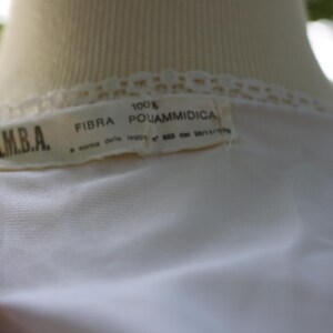 May include: A white garment featuring a lace trim along the neckline and a rectangular fabric label. The label displays the text I.M.B.A., 100% FIBRA POLIAMMIDICA, and additional text referencing a law from 1973. The fabric appears smooth and lightweight.
