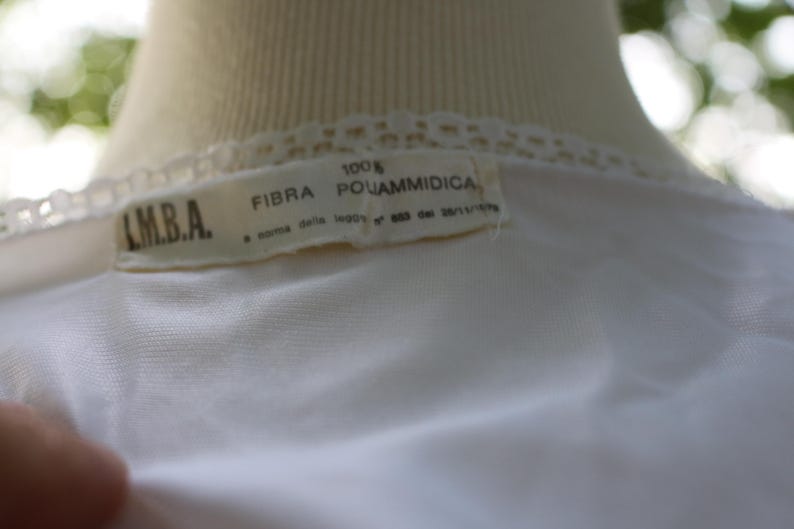 May include: A white garment featuring a lace trim along the neckline and a rectangular fabric label. The label displays the text I.M.B.A., 100% FIBRA POLIAMMIDICA, and additional text referencing a law from 1973. The fabric appears smooth and lightweight.