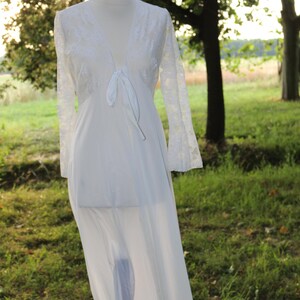 May include: A long white robe featuring sheer lace long sleeves and a lace bodice with a deep V-neckline. The garment includes a front tie closure at the chest and a flowing, semi-sheer skirt that extends to the floor, displayed on a mannequin.