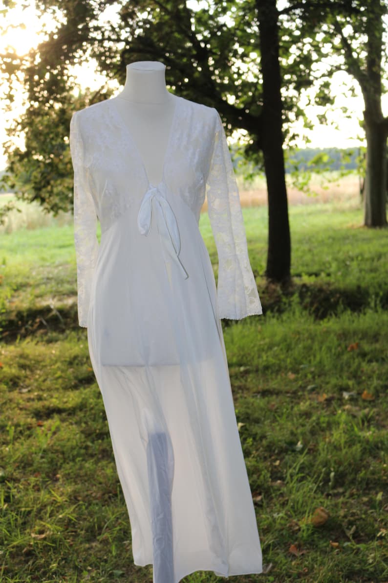 May include: A long white robe featuring sheer lace long sleeves and a lace bodice with a deep V-neckline. The garment includes a front tie closure at the chest and a flowing, semi-sheer skirt that extends to the floor, displayed on a mannequin.