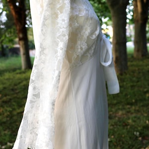 May include: A white dress featuring long, sheer lace sleeves with a floral pattern. The bodice is constructed from matching white lace, while the skirt is made of a solid, opaque white fabric. A white fabric tie is visible at the side waist.