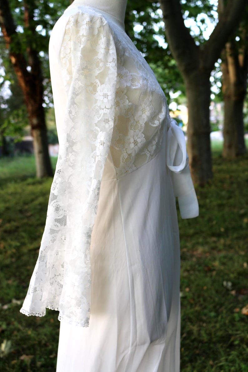 May include: A white dress featuring long, sheer lace sleeves with a floral pattern. The bodice is constructed from matching white lace, while the skirt is made of a solid, opaque white fabric. A white fabric tie is visible at the side waist.