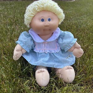 May include: A doll with light skin, blue eyes, and curly yellow yarn hair. The doll wears a light blue dress with white polka dots, a white collar with eyelet lace trim, and a pink ribbon detail at the center of the chest. The doll has soft limbs.