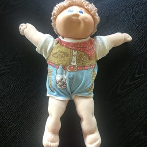 Könnte beinhalten: Eine Vintage-Cabbage-Patch-Kid-Puppe mit einem lächelnden Gesicht, blauen Augen und hellbraunem, lockigem Haar. Die Puppe trägt ein weißes Hemd, eine Weste mit rotem Bandana und blaue Shorts. Die Arme der Puppe sind ausgestreckt.