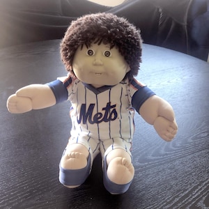 May include: A Cabbage Patch Kids doll wearing a white and blue pinstripe baseball uniform with the word "Mets" across the chest. The doll has brown, curly hair and is seated on a dark surface.