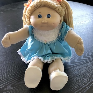 Puede incluir: Una muñeca vintage con cabello rubio de hilo en coletas, aseguradas con lazos naranjas. La muñeca tiene ojos azules y lleva un vestido azul claro con ribete de encaje blanco y zapatos blancos. La muñeca está sentada sobre una superficie oscura.