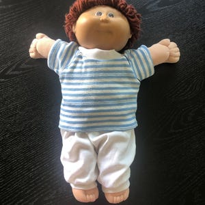 Könnte beinhalten: Eine Vintage-Puppe mit rundem Gesicht, braunen Haaren und blauen Augen. Die Puppe trägt ein blau-weiß gestreiftes Oberteil und eine weiße Hose. Die Arme der Puppe sind ausgestreckt.