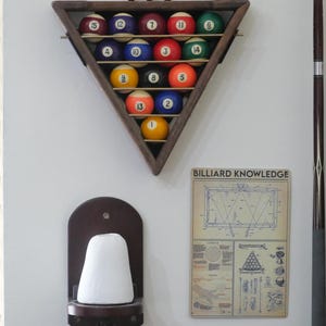 May include: A triangular wooden wall rack holding fifteen numbered billiard balls in a rack formation. Below it, a dark wood wall-mounted holder with a white chalk cone and a rectangular sign titled BILLIARD KNOWLEDGE with diagrams.
