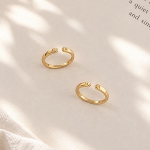 Peut inclure: Deux bagues ouvertes de couleur dorée avec une finition lisse et polie. Chaque bague présente deux lettres majuscules gravées aux extrémités de l'anneau. Une bague affiche les lettres E et K, tandis que l'autre affiche les lettres M et A.