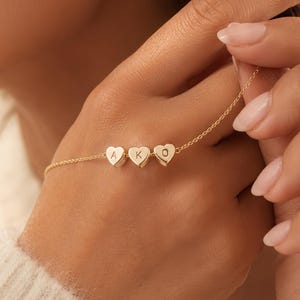 Op de afbeelding: Een delicate gouden kettingarmband met drie kleine hartvormige bedels. Elk hartje is gegraveerd met een enkele letter, namelijk de letters A, K en O, gerangschikt in een horizontale rij langs de dunne, gepolijste goudkleurige metalen ketting.