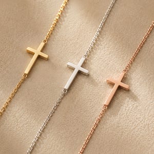 Könnte beinhalten: Drei zarte Armbänder mit einem kleinen, mittig platzierten Kreuzanhänger. Das Set besteht aus einer goldfarbenen, einer silberfarbenen und einer roségoldfarbenen Kette, die jeweils aus feinen, polierten Metallgliedern bestehen.