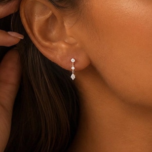 May include: A delicate drop earring featuring three small, clear, round-cut stones set in a vertical line on a thin metal post. The earring hangs from the earlobe, showcasing a minimalist design with a polished finish and secure backing.