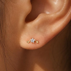 May include: A gold-toned ear stud featuring a central star shape set with a clear, sparkling stone, flanked by two smaller plain gold-toned stars on either side. The earring is positioned on the earlobe, showcasing a delicate and celestial design.