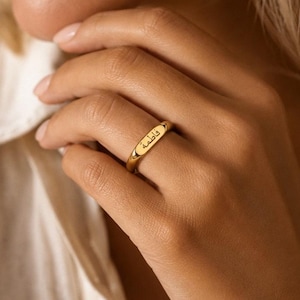 Peut inclure: Une bague dorée dotée d'une face ovale lisse et plate, gravée du prénom arabe Fatima. L'anneau est poli et fin, entourant le doigt avec un design épuré qui met en valeur l'inscription en lettres cursives sombres et bien lisibles.