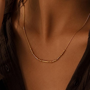 Puede incluir: Un delicado collar de cadena dorada que presenta una serie de pequeñas cuentas cilíndricas y diminutos acentos esféricos espaciados a lo largo de la parte inferior de la cadena. El collar descansa sobre el cuello, mostrando un acabado metálico.