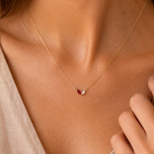 Peut inclure: Un collier à fine chaîne dorée orné de deux petites pierres précieuses en forme de goutte d'eau placées au centre. L'une des pierres est d'un rouge profond, tandis que l'autre est transparente et incolore. Le collier repose sur le cou.