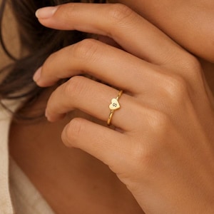 Puede incluir: Un anillo fino de color dorado que presenta un pequeño dije en forma de corazón centrado en la banda. El corazón está grabado con la letra mayúscula S en una fuente oscura y clara. El anillo tiene un acabado metálico pulido y un diseño fino.