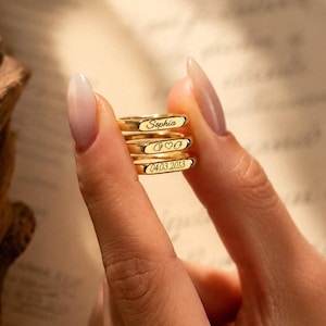 May include: Three stacked gold rings with flat rectangular faces. The top ring is engraved with the name Sophia, the middle ring features a heart symbol between two letters, and the bottom ring displays the numerical date 04.03.2013.