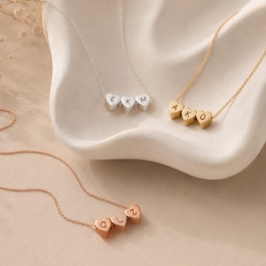 May include: Three delicate necklaces featuring heart-shaped charms. One silver necklace with charms engraved with E, K, M. One gold necklace with charms engraved with A, K, O. One rose gold necklace with charms engraved with Q, L, Z. Each chain is thin.