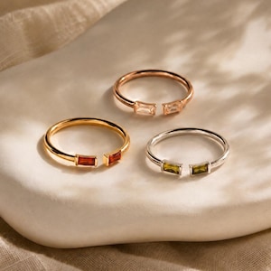 May include: Three open-style rings featuring baguette-cut gemstones. One ring is rose gold with clear stones, one is gold with orange-red stones, and one is silver with olive-green stones. Each ring has a thin band and two rectangular stones.