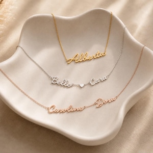 May include: Three delicate necklaces with cursive name pendants. The top gold necklace features the name Alberta. The middle silver necklace displays the names Belle and Cara separated by a small heart. The bottom rose gold necklace shows Caroline and Dora.