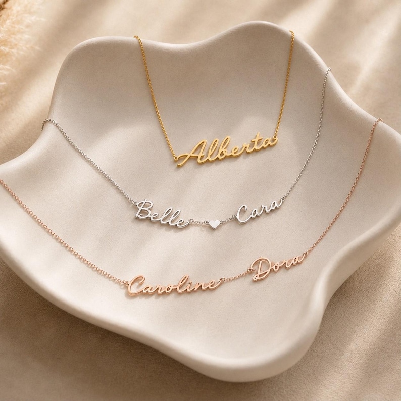 May include: Three delicate necklaces with cursive name pendants. The top gold necklace features the name Alberta. The middle silver necklace displays the names Belle and Cara separated by a small heart. The bottom rose gold necklace shows Caroline and Dora.