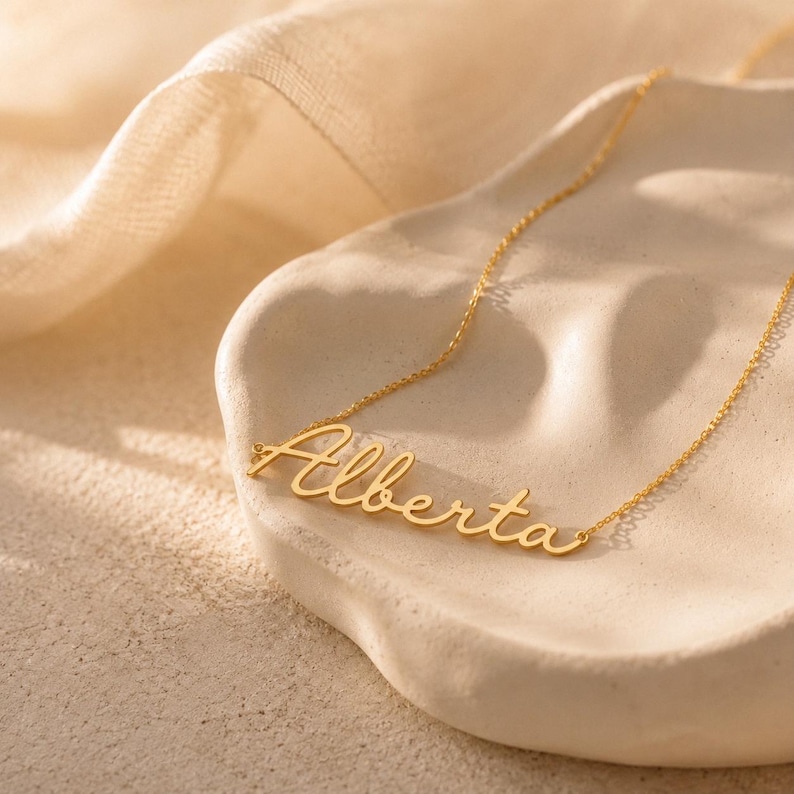 May include: A gold-toned metal necklace featuring a delicate chain and a nameplate pendant with the name Alberta written in a cursive script font. The necklace is displayed on a textured, off-white, organic-shaped surface.