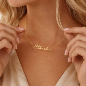 May include: A gold-toned necklace featuring a delicate chain and a central nameplate pendant displaying the name Alberta in a cursive, flowing script font. The metal has a polished finish that reflects light, creating a bright, metallic appearance.