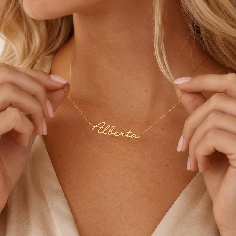 May include: A gold-toned necklace featuring a delicate chain and a central nameplate pendant displaying the name Alberta in a cursive, flowing script font. The metal has a polished finish that reflects light, creating a bright, metallic appearance.