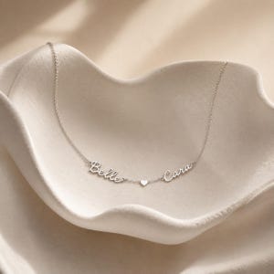 May include: A silver-toned necklace featuring a delicate chain with two script-style name pendants reading Belle and Cara, separated by a small, centered heart charm. The metal has a polished finish and the names are connected directly to the chain.