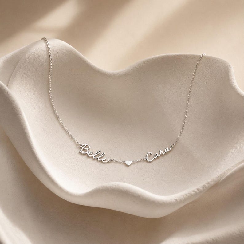 May include: A silver-toned necklace featuring a delicate chain with two script-style name pendants reading Belle and Cara, separated by a small, centered heart charm. The metal has a polished finish and the names are connected directly to the chain.