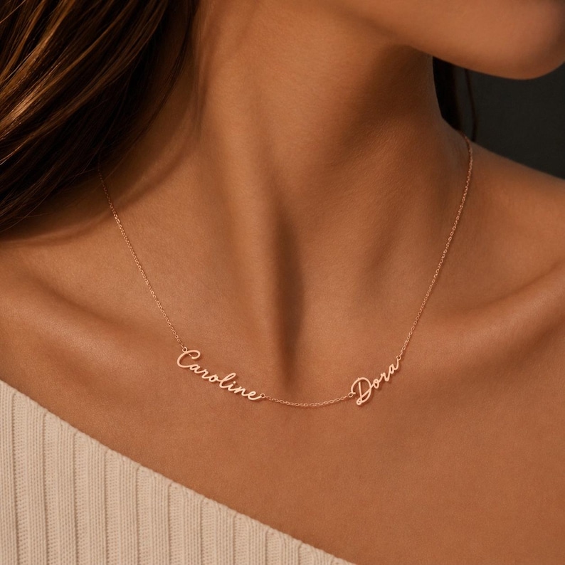 May include: A rose gold-toned necklace featuring two names, Caroline and Dora, written in a delicate cursive script. The names are integrated into a thin, fine chain that rests against the collarbone, creating a personalized jewelry piece.