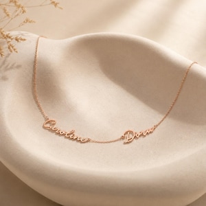 May include: A rose gold-toned metal necklace featuring two names, Caroline and Dora, written in a cursive script font. The delicate chain connects the two names, creating a continuous piece of jewelry with a polished, metallic finish.