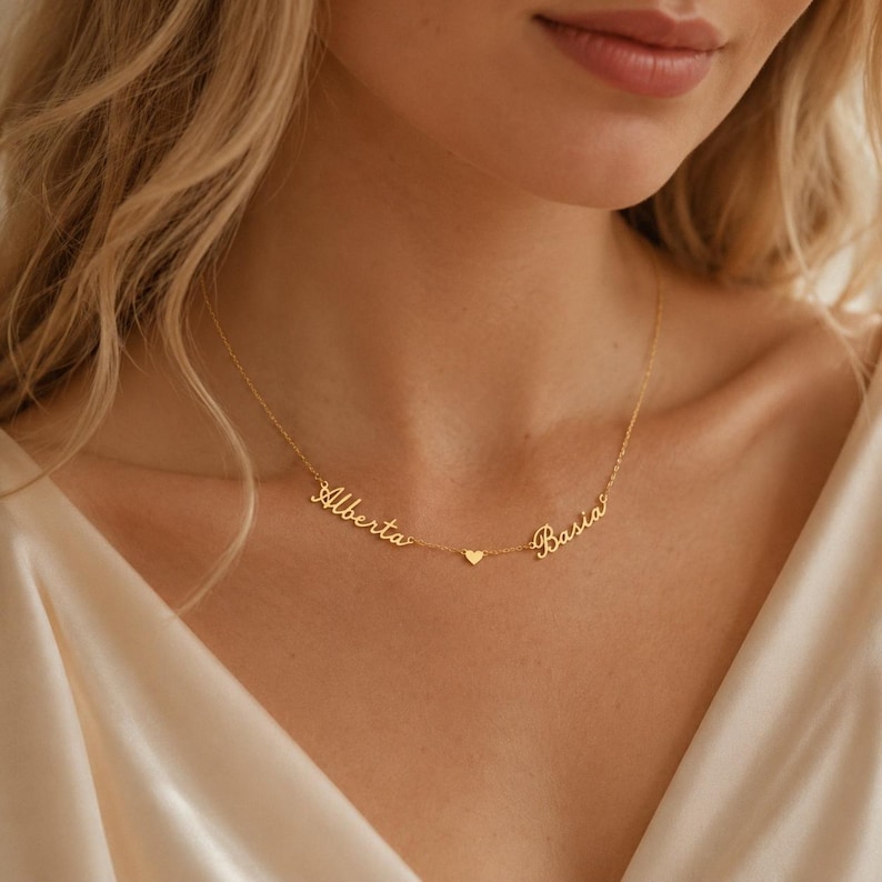 May include: A gold-toned necklace featuring two names written in a cursive script, Alberta and Basia, connected by a delicate chain. A small, solid gold-toned heart charm is positioned centrally between the two names on the thin metal chain.