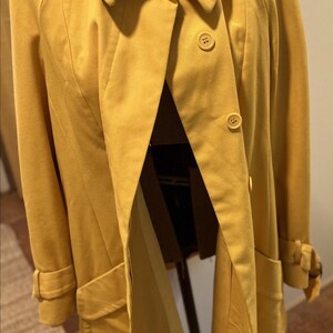 May include: A mustard yellow coat featuring a wide collar, long sleeves with buckled cuffs, and a front button closure. The garment has two front pockets and is displayed on a mannequin, showcasing a structured design with visible seams.
