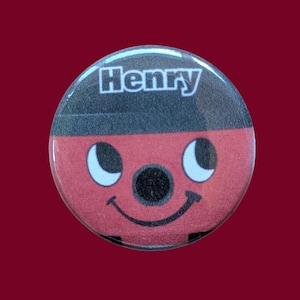 May include: A circular pinback button featuring a red face with a black top section, large white eyes, a black circular nose, and a black smiling mouth. The name Henry is printed in white capital letters across the top black section.
