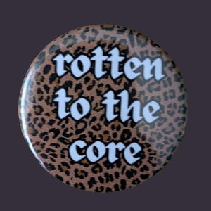 May include: A circular pin-back button featuring a brown and black leopard print pattern. The text "rotten to the core" is printed in white, bold, sans-serif capital letters across the centre of the button, covering the animal print background.