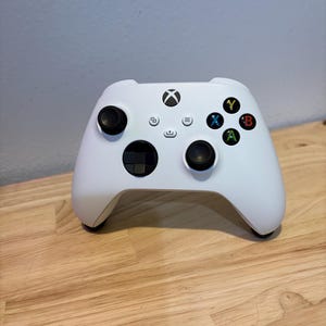 以下が含まれることがあります： 黒いサムスティックとボタンが付いた白いXboxコントローラー。 コントローラーにはXboxのロゴと、Y（黄色）、X（青）、B（赤）、A（緑）のカラーボタンがあります。 木製の表面に置かれています。