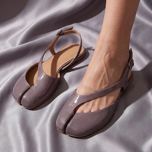 Op de afbeelding: Een paar glanzende, lavendelkleurige split-toe flats met verstelbare enkelbandjes. De schoenen hebben een uniek ontwerp, met een apart compartiment voor de grote teen. De flats staan tegen een satijnen achtergrond.