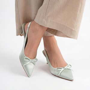 May include: Pale green satin slingback heels with pointed toes and small bows. The shoes have a delicate, feminine design and a low heel. The background is white.