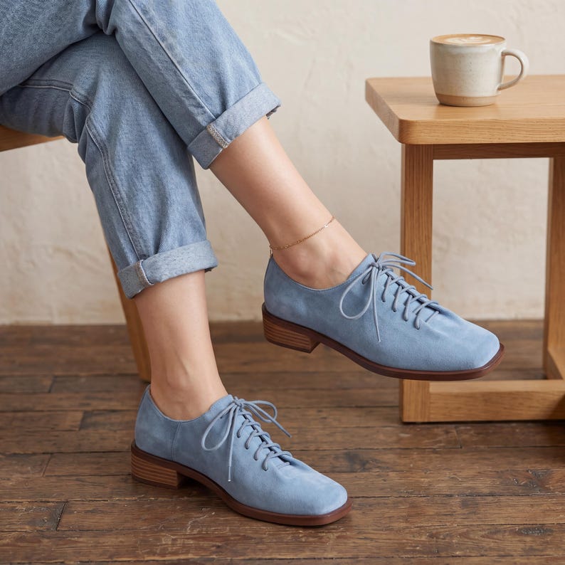 May include: Light blue suede lace-up shoes with a square toe and a low wooden heel. The shoes are worn with light blue jeans. The laces are light blue and the soles are brown. The shoes are a classic style.