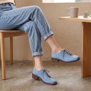 May include: Light blue suede oxford shoes with brown heels and laces. The shoes are worn by a person wearing blue jeans. The shoes have a classic design and a low heel.